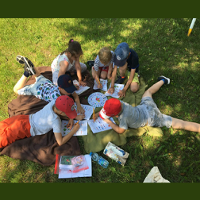 Grupa dzieci siedzi na kocu na trawie i rysuje kolorowymi kredkami na kartkach.