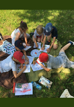 Grupa dzieci siedzi na kocu na trawie i rysuje kolorowymi kredkami na kartkach.