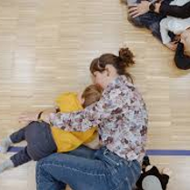 Na zdjęciu fragment warsztatów dla dzieci. Na podłodze leżą dorośli przytulając dzieci.