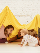 Na zdjęciu fragment warsztatów dla dzieci. Kobieta wraz z kilkorgiem dzieci siedzi na podłodze nakrywając ich żółtym materiałem. Wszyscy są uśmiechnięci.