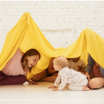 Na zdjęciu fragment warsztatów dla dzieci. Kobieta wraz z kilkorgiem dzieci siedzi na podłodze nakrywając ich żółtym materiałem. Wszyscy są uśmiechnięci.