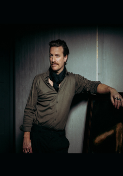 Na zdjęciu Parov Stelar opiera się o czarny ekran i patrzy w obiektyw.