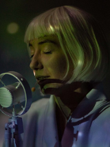 na zdjęciu twarz kobiety o jasnych włosach, z grzywką, przed nią mikrofon. Kobieta ma zamknięte oczy.