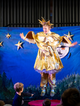 Kadr ze spektaklu. Kobieta ubrana w złoty strój gwiazdki wisi zawieszona na tle niebieskiej płachty symbolizującej niebo. Przed sceną widać chłopca i głopwy kilku innych osób.