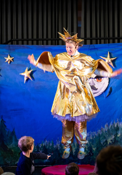 Kadr ze spektaklu. Kobieta ubrana w złoty strój gwiazdki wisi zawieszona na tle niebieskiej płachty symbolizującej niebo. Przed sceną widać chłopca i głopwy kilku innych osób.