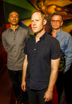 Członkowie zespołu pozują do zdjęcia w zaciemnionych wnętrzach klubu. Za nimi bordowa kanapa narożnikowa i stolik.