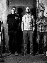 Czarno-białe zdjęcie, na którym widać członków tria pozujących na tle drzwi.