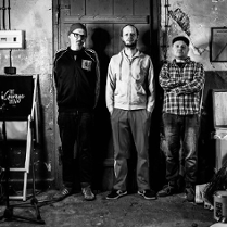 Czarno-białe zdjęcie, na którym widać członków tria pozujących na tle drzwi.