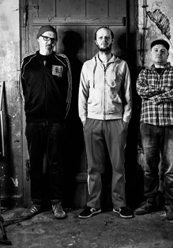 Czarno-białe zdjęcie, na którym widać członków tria pozujących na tle drzwi.