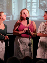 Zdjęcie ze spektaklu. Trzy aktorki w trakcie występu stoją przy mikrofonie.