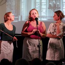 Zdjęcie ze spektaklu. Trzy aktorki w trakcie występu stoją przy mikrofonie.