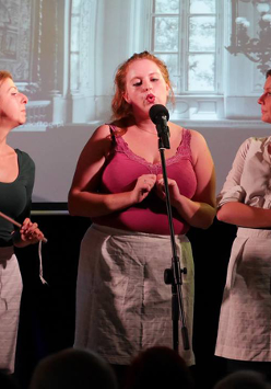 Zdjęcie ze spektaklu. Trzy aktorki w trakcie występu stoją przy mikrofonie.