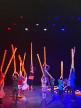 Na zdjęciu dwoje aktorów oraz z dziećmi stoją na scenie trzymając nad głowami długie tuby przypominające szyje żyraf