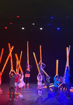 Na zdjęciu dwoje aktorów oraz z dziećmi stoją na scenie trzymając nad głowami długie tuby przypominające szyje żyraf