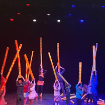 Na zdjęciu dwoje aktorów oraz z dziećmi stoją na scenie trzymając nad głowami długie tuby przypominające szyje żyraf