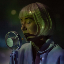 na zdjęciu twarz kobiety o jasnych włosach, z grzywką, przed nią mikrofon. Kobieta ma zamknięte oczy.