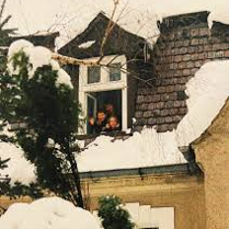 Na zdjęciu widać ośnieżony dach domu z małym wysuniętym oknem mansardowym. W otwartym oknie stoją trzy uśmiechnięte dzieci, które wyglądają na zadowolone i machają w stronę fotografa. Dach pokryty jest grubą warstwą śniegu, a gałęzie drzew wokół również są przysypane. Scena sprawia wrażenie zimowego, rodzinnego i radosnego momentu.