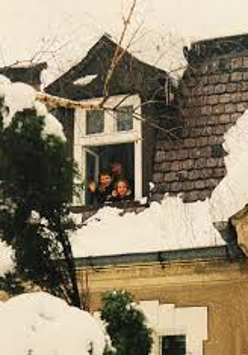 Na zdjęciu widać ośnieżony dach domu z małym wysuniętym oknem mansardowym. W otwartym oknie stoją trzy uśmiechnięte dzieci, które wyglądają na zadowolone i machają w stronę fotografa. Dach pokryty jest grubą warstwą śniegu, a gałęzie drzew wokół również są przysypane. Scena sprawia wrażenie zimowego, rodzinnego i radosnego momentu.