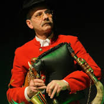 Na zdjęciu dudziarz w czerwonym ubraniu trzymający instrument muzyczny.