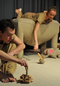 Zdjęcie aktorów w trakcie spektaklu. Trzech aktorów ubranych na beżowo z młynkowymi instrumentami. Beżowy słonik z różowymi uszami.