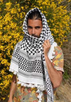 Grafika na tle miętowego koloru. Na środku znajduje się zdjęcie mężczyzny z brodą i długimi ciemnymi włosami, ubranego w kefiję.