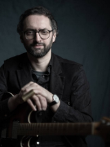 Mężczyzna w ciemnym stroju, trzymający skrzyżowane ręce na gitarze przed sobą. Ciemne tło.