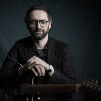 Mężczyzna w ciemnym stroju, trzymający skrzyżowane ręce na gitarze przed sobą. Ciemne tło.