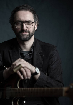 Mężczyzna w ciemnym stroju, trzymający skrzyżowane ręce na gitarze przed sobą. Ciemne tło.