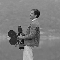 Czarno-białe zdjęcie mężczyzny stojacego bokiem do obiektywu i trzymającego kamerę na tle zamglonego krajobrazu.