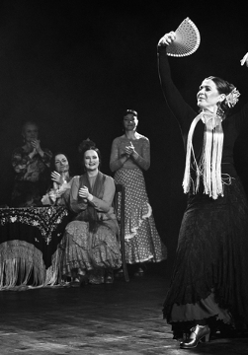 Czarno-białe zdjęcie tancerki flamenco w tanecznej pozie z wachlarzem. Na drugim placnie kilka kobiet obserwujących występ i klaszczących w dłonie.