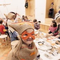 Uśmiechnięta dziewczynka w przebraniu z maską na twarzy. W tle dzieci siedzące przy stole przy pracach plastycznych.