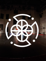 Zdjęcie sceny i czterech stojących na niej osób. Na pierwszym planie biała grafika: cztery połaczone ze sobą kółka, otoczone przerywaną obwódką.