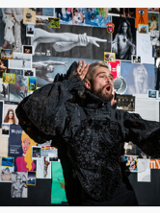 Na zdjęciu mężczyzna ubrany w czarny strój, gestykuluje i mówi lub śpiewa. W tle kolorowa ściana wyklejona zdjęciami, wycinkami i obrazkami.