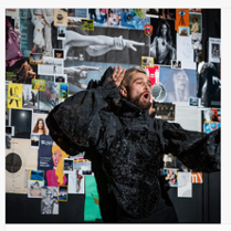 Na zdjęciu mężczyzna ubrany w czarny strój, gestykuluje i mówi lub śpiewa. W tle kolorowa ściana wyklejona zdjęciami, wycinkami i obrazkami.