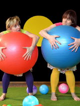 Dwie kolorowo ubrane kobiety, stojące boso na scenie, jedna z nich trzyma przed sobą dużą czerwoną piłkę, druga niebieską.