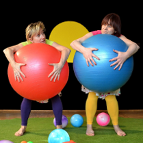 Dwie kolorowo ubrane kobiety, stojące boso na scenie, jedna z nich trzyma przed sobą dużą czerwoną piłkę, druga niebieską.