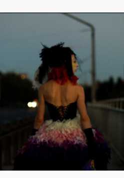 na zdjęciu widoczna postać kobiety idąca wieczorem chodnikiem ulicy, ubrana jest w sukienkę z gorsetem, z odsłoniętymi ramionami, na głowie ma woalkę z czerwonymi i czarnymi piórami i wstążkami.