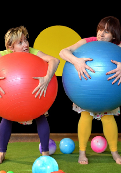 Dwie kolorowo ubrane kobiety, stojące boso na scenie, jedna z nich trzyma przed sobą dużą czerwoną piłkę, druga niebieską.