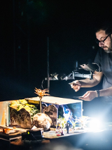 Mężczzyna przy oświetlonym stole, na którym znajdują się różne przedmiety. Mężczyzna trzyma w rękach dwie karty, na które patrzy.