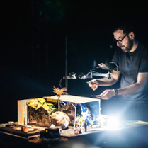 Mężczzyna przy oświetlonym stole, na którym znajdują się różne przedmiety. Mężczyzna trzyma w rękach dwie karty, na które patrzy.