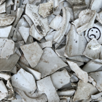 Fragmenty potłuczonych białych, brudnych naczyń; na jednym kawałku naczynia narysowana smutna buźka.