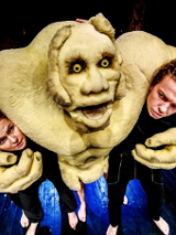 Bajkowa postać ze spektaklu, obejmująca ramionami dwóch aktorów, za nią kolejne dwie osoby.