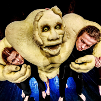 Bajkowa postać ze spektaklu, obejmująca ramionami dwóch aktorów, za nią kolejne dwie osoby.