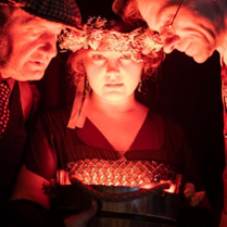 Oświetlone twarze dwóch mężczyzn i kobiety w wianku na głowie. Twarze trojga ludzi są blisko siebie, patrzą na coś w dole.