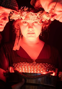 Oświetlone twarze dwóch mężczyzn i kobiety w wianku na głowie. Twarze trojga ludzi są blisko siebie, patrzą na coś w dole.