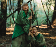 Dwie kobiety w lesie, ubrane w zielone atroje, trzymają cienki pień drzewa.