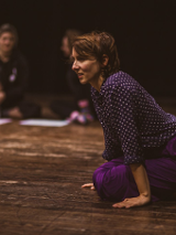 Na podłodze klęcząca kobieta, podpierająca się rękami. W tle dwie osoby, siedzące na podłodze z podkurczonymi nogami.