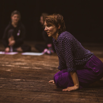 Na podłodze klęcząca kobieta, podpierająca się rękami. W tle dwie osoby, siedzące na podłodze z podkurczonymi nogami.