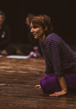 Na podłodze klęcząca kobieta, podpierająca się rękami. W tle dwie osoby, siedzące na podłodze z podkurczonymi nogami.