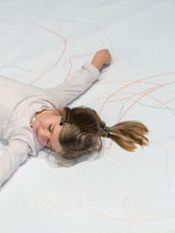 Zdjęcie dziewczynki leżącej z rozłożonymi rękami na dużym arkuszu papieru popisanego kredkami.